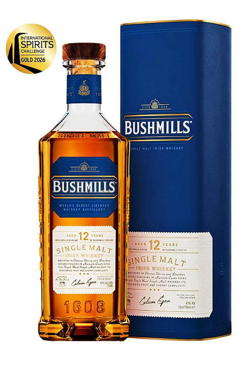A bottle of Bushmills 12 Years Single Malt Irish Whiskey next to its blue cylindrical packaging box, with an International Spirits Challenge Gold 2026 seal in the top-left corner.