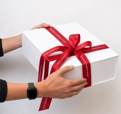 A white rectangular gift box being held by two hands, tied with a wide red satin ribbon and bow. The ribbon has white text that reads 'whiskerybites.