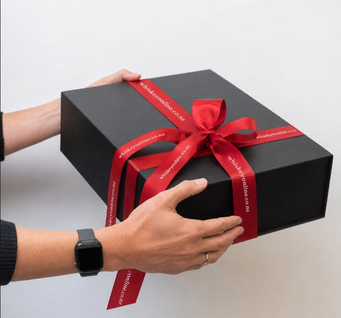 A black rectangular gift box tied with a red satin ribbon and bow, held by two hands against a plain light background. The ribbon has white text reading 'whitakerwooden.